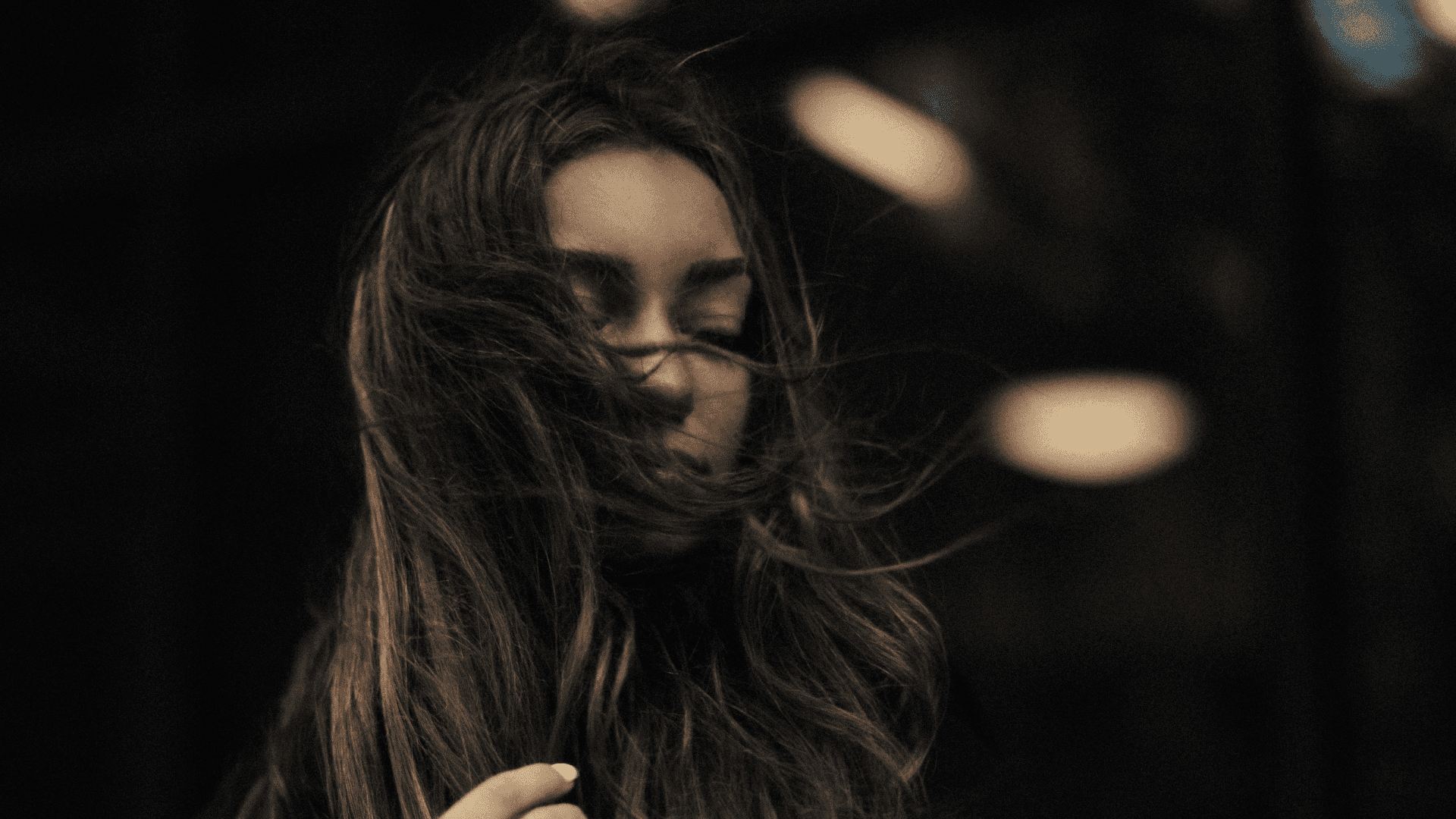 Young woman with long hair blown by wind, eyes closed, in a dark, moody background.